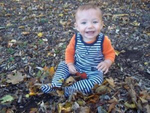 A young toddler in an orange shirt and striped overalls sits in a pile of leaves at Glendale Heights Child Care in Glendale, WI