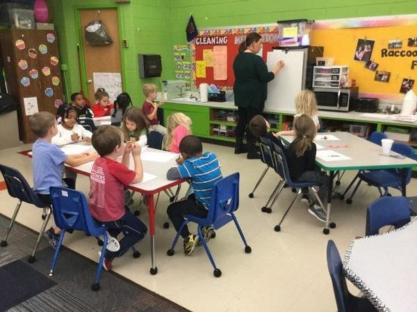 Children sitting at a white tables in a classroom while a teacher uses a whiteboard at the front of a classroom in Glendale Heights Child Care in Glendale, WI