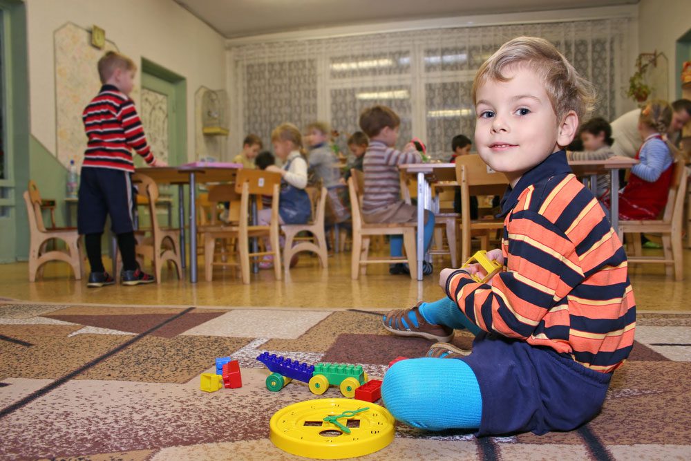 A young boy sitting on the carpeted floor with toys as other children sit at wooden tables
